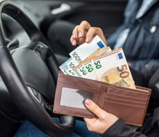 Eine unkenntliche weibliche Person sitzt in ihrem Auto hinter dem Steuer. Im Hintergrund sitzt ein männlicher Beifahrer. Die weibliche Hand zieht 50-Euro- und 100-Euro-Scheine aus ihrem Geldbeutel, um immer höhere Bußgelder zu bezahlen.