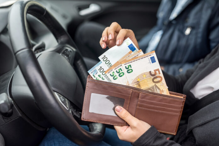 Eine unkenntliche weibliche Person sitzt in ihrem Auto hinter dem Steuer. Im Hintergrund sitzt ein männlicher Beifahrer. Die weibliche Hand zieht 50-Euro- und 100-Euro-Scheine aus ihrem Geldbeutel, um immer höhere Bußgelder zu bezahlen.