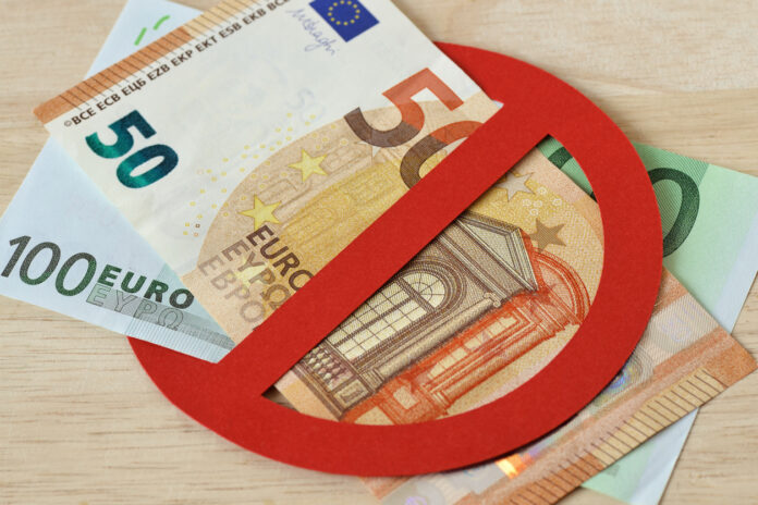 Auf einem Tisch aus hellem Holz liegen verschiedene Euro-Scheine. Zu sehen sind ein 50- und ein 100-Euro-Schein. Diese sind in ein ausgeschnittenes rotes Verbotsschild gesteckt. Dies soll andeuten, dass Bargeld hier wohl verboten ist.