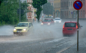 Mehrere Autos fahren über eine nasse Straße. Es regnet stark. Vorne sieht man ein Straßenschild.