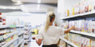 Eine Frau steht zwischen zwei Regalen im Supermarkt. Sie hält einen Einkaufskorb.