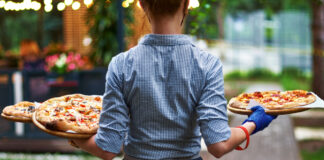 Eine Kellnerin trägt mehrere Pizzen draußen in einem Biergarten zum Tisch