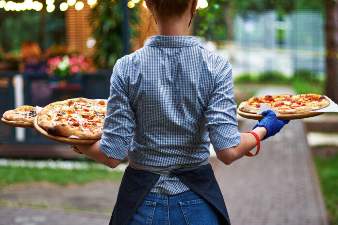 Eine Kellnerin trägt mehrere Pizzen draußen in einem Biergarten zum Tisch