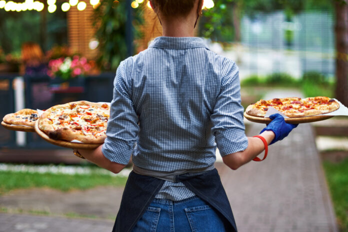 Eine Kellnerin trägt mehrere Pizzen draußen in einem Biergarten zu einem Tisch. Im Hintergrund stehen mehrere Tische und eine Lichterkette beleuchtet das Lokal von außen.
