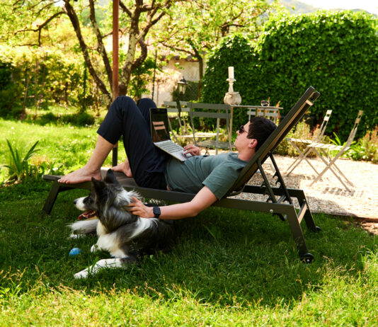 Ein Mann liegt auf einer Liege im Garten und hat einen Laptop auf dem Schoß. Ein Hund liegt neben ihm im Gras.