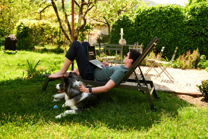 Ein Mann liegt auf einer Liege im Garten und hat einen Laptop auf dem Schoß. Ein Hund liegt neben ihm im Gras.