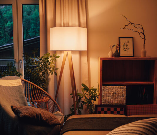 Verschiedene Gegenstände und Möbel stehen in einem Haushalt. Ein Bild steht auf einem Regal, ein gemütlicher Stuhl und eine Couch schmücken das Wohnzimmer, in dem das warme Licht einer Lampe im brennt.