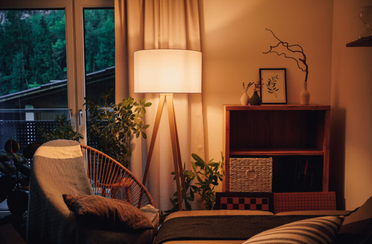 Verschiedene Gegenstände und Möbel stehen in einem Haushalt. Ein Bild steht auf einem Regal, ein gemütlicher Stuhl und eine Couch schmücken das Wohnzimmer, in dem das warme Licht einer Lampe im brennt.
