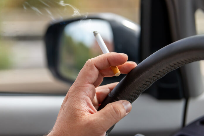 Ein Mann hält in der Hand, mit der er das Lenkrad seines Autos bedient, eine brennende Zigarette.