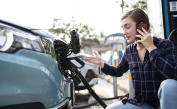 Eine Frau hockt neben ihrem Auto mit einem Handy und telefoniert, dabei betankt sie den Wagen
