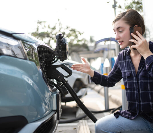 Eine Frau hockt neben ihrem Auto mit einem Handy und telefoniert, dabei betankt sie den Wagen