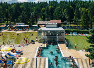 Ein großer Badepark mit vielen Leuten, die schwimmen, planschen, tauchen und Spaß haben. Neben einem Freibad gibt es große Wiesenflächen, auf denen die Menschen liegen oder picknicken können.