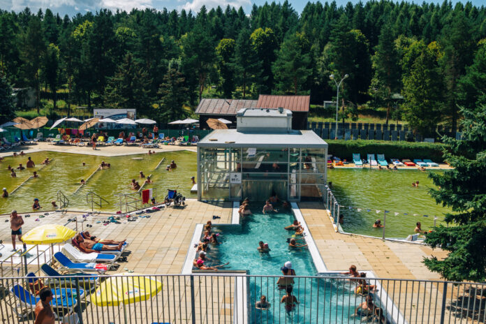 Ein großer Badepark mit vielen Leuten, die schwimmen, planschen, tauchen und Spaß haben. Neben einem Freibad gibt es große Wiesenflächen, auf denen die Menschen liegen oder picknicken können.