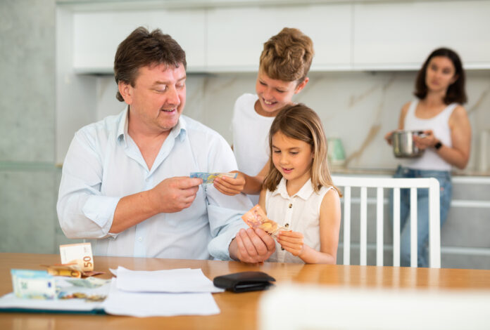Ein Vater zeigt seinem Sohn und seiner Tochter Geldscheine und Münzen an einem Tisch. Im Hintergrund steht die Mutter mit einem Topf in der Hand in der Küche.