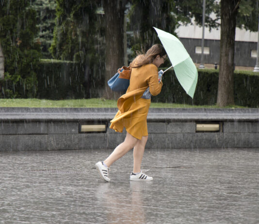 Junge Frau im gelben Kleid mit einem Regenschirm im strömenden Regen läuft über einen Platz