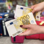 Eine Frau hält Euro Scheine in der Hand. Ihre Fingernägel sind rot lackiert, während im Hintergrund einige Koffer und unterschiedliches Gepäck steht.