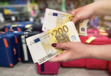 Eine Frau hält Euro Scheine in der Hand. Ihre Fingernägel sind rot lackiert, während im Hintergrund einige Koffer und unterschiedliches Gepäck steht.