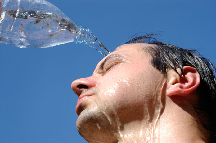 Nahaufnahme eines Mannes, der sich eine Flasche voller Wasser über den Kopf gießt. Ihm ist offensichtlich sehr warm. Im Hintergrund erkennt man einen strahlend blauen Himmel und die Sonne prallt herab. Es scheint Sommer und damit sehr heiß zu sein. Der Mann schwitzt und ist auf der Suche nach einer Abkühlung.