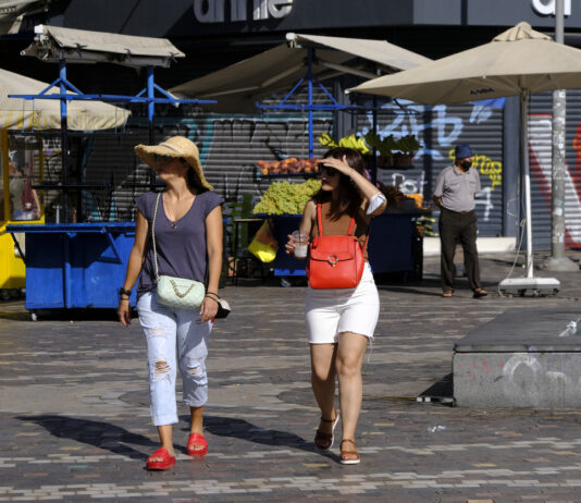 Menschen gehen während einer Hitzewelle durch die Straße in einem Stadtzentrum. Sie tragen ein Cap um sich vor der Sonne zu schützen und Shorts.