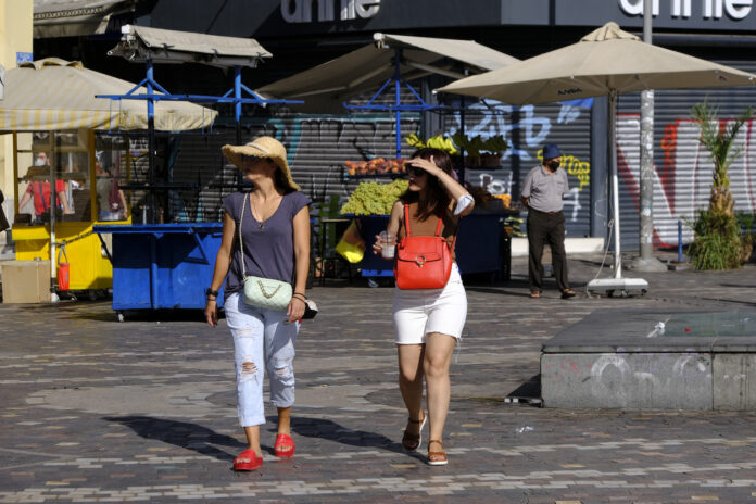 Menschen gehen während einer Hitzewelle durch die Straße in einem Stadtzentrum. Sie tragen ein Cap um sich vor der Sonne zu schützen und Shorts.