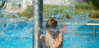 Ein Junge ist in einem Freibad und kühlt sich unter einer Fontäne ab. Er hat sichtlich Freude an dem Wasserspiel, während um ihn herum das Wasser in Bewegung ist. Der Pool strahlt in einem türkisen Blau.