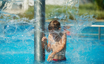 Ein Junge ist in einem Freibad und kühlt sich unter einer Fontäne ab. Er hat sichtlich Freude an dem Wasserspiel, während um ihn herum das Wasser in Bewegung ist. Der Pool strahlt in einem türkisen Blau.