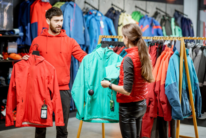Ein Verkäufer übergibt einer Kundin im Laden zwei verschiedene Jacken zur Anprobe
