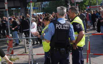 Polizisten und Mitarbeiter des Ordnungsamtes überwachen eine Demonstration oder eine Menschenansammlung auf der Straße. Sie tragen Warnwesten mit der Aufschrift "Polizei". Außerdem sind sie bewaffnet.