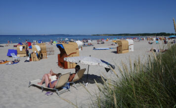 Viele Urlauber entspannen an einem weißen Sandstrand vor einem kristallklaren, blauem Meer vor blauem Himmel auf Decken und in diversen Strandkörben, die an der Küste verstreut sind und Schutz vor der Sonne bieten.