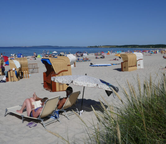 Viele Urlauber entspannen an einem weißen Sandstrand vor einem kristallklaren, blauem Meer vor blauem Himmel auf Decken und in diversen Strandkörben, die an der Küste verstreut sind und Schutz vor der Sonne bieten.
