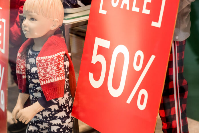 Die Nahaufnahme eines Schaufensters. Im Fenster ist ein rotes Rabattschild mit der Aufschrift "Sale" und "50%" zu sehen. Daneben steht eine kleine Schaufensterpuppe in einem Schlafanzug und mit einem roten Pullover um die Schultern herum.