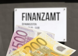 Ein 200- und ein 500-Euro-Geldschein werden in die Kamera gehalten. Im Hintergrund befindet sich an einem Gebäude ein weißes Schild mit der Aufschrift "Finanzamt". Die darunter befindliche Schrift ist nicht zu erkennen, da sie verschwommen ist.