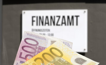 Ein 200- und ein 500-Euro-Geldschein werden in die Kamera gehalten. Im Hintergrund befindet sich an einem Gebäude ein weißes Schild mit der Aufschrift "Finanzamt". Die darunter befindliche Schrift ist nicht zu erkennen, da sie verschwommen ist.