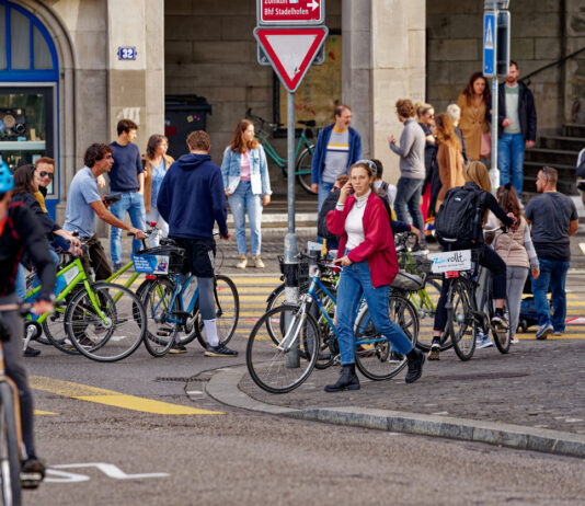 Mehrere Radfahrer überqueren eine Straße zu Fuß, sie schieben ihr Fahrrad. Im Hintergrund stehen einige Fußgänger am Straßenrand und schauen sich um.