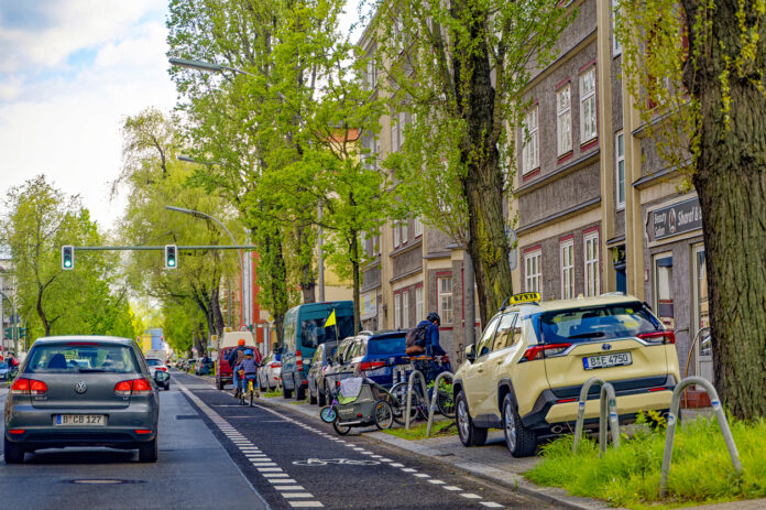 Straßenzene mit parkenden Autos, Fahrradweg mit Fahrradfahrern und Straße in einem Wohngebiet Berlins.