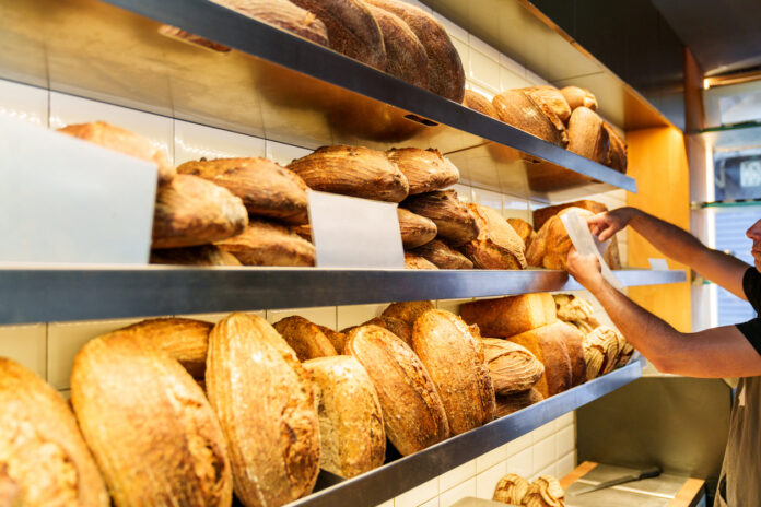 Ein Regal in der Bäckerei mit frischen Backwaren. Auf mehreren Etagen findet man verschiedene Sorten von Brot und einige Panini. Ein Mitarbeiter befindet sich in der Bäckerei und bringt ein Schild an.