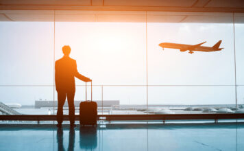 Eine Person steht mit einem Koffer am Flughafen und schaut einem Flugzeug nach, das in den Urlaub startet.