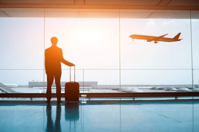 Eine Person steht mit einem Koffer am Flughafen und schaut einem Flugzeug nach, das in den Urlaub startet.