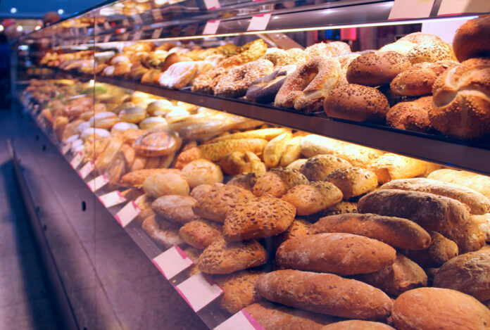 Blick auf verschiedene Backwaren in einer Bäckerei. Die Backwaren legen nebeneinander in einer Vitrine. Unter anderem sind Brötchen und süßes Gebäck zu finden. Weiße Schilder weisen auf die Produkte und ihren Preis hin.