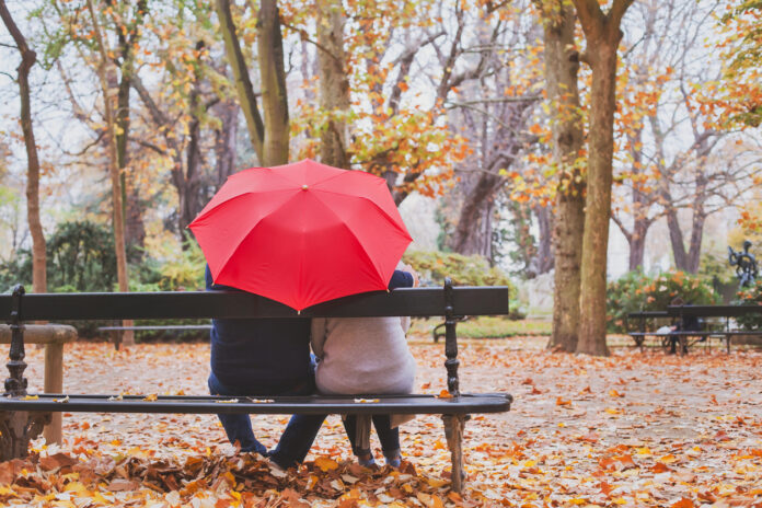 Ein Paar sitzt unter dem Regenschirm auf einer Bank im Herbstlaub.