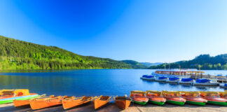 Zu sehen ist der Titisee mit seinem strahlend blauen Wasser und dem sauberen Strand. Im Hintergrund sind Berge zu sehen.