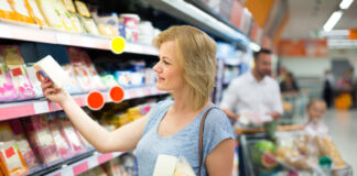 Eine Frau steht im Supermarkt an einem Kühlregal mit Käse. Dahinter steht ein Mann mit einem Kind im Einkaufswagen.