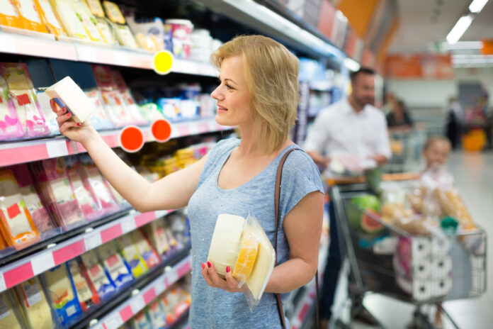Eine Frau steht im Supermarkt an einem Kühlregal mit Käse. Dahinter steht ein Mann mit einem Kind im Einkaufswagen.