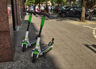 Zwei weiß-grüne E-Scooter parken am Rande einer Einkaufsstraße. Im Hintergrund parken ein paar Autos, und Fußgänger gehen ihren Weg. Es soll ein neues Verbot für E-Roller kommen.
