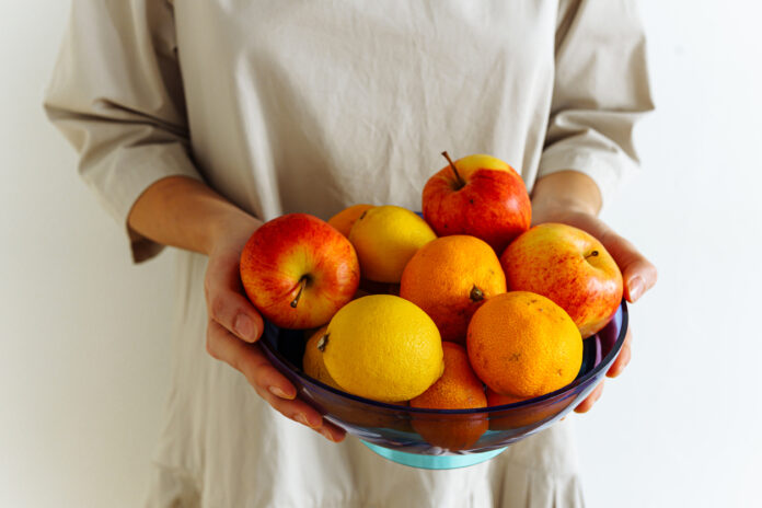 Eine Schale mit Obst. Es sind Äpfel, Clementinen und Zitronen zu sehen. Die Schale wird von beiden Händen einer Frau gehalten, deren Oberkörper im Bild zu sehen ist. Die Frau trägt ein helles Oberteil.