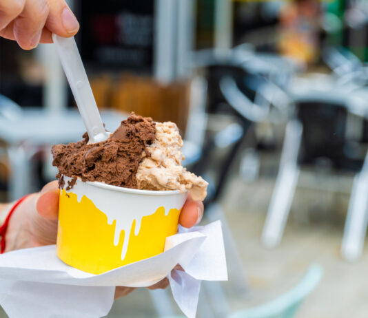 Ein Eisbecher wird gegessen. Eine Person sitzt in einem Eiscafé und hält einen großen gelben und weißen Eisbecher in der Hand, in dem sich Eiskugeln verschiedener Geschmacksrichtungen türmen.