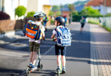 Zwei Schulkinder mit Schulranzen und Rollern auf dem Weg zur Schule.