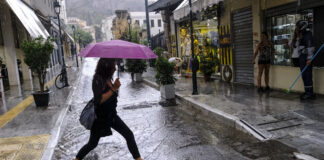 Frau mit Regenschirm überquert Straße im Regen. Im Hintergrund sieht man mehrere Geschäfte die hell erleuchtet sind.