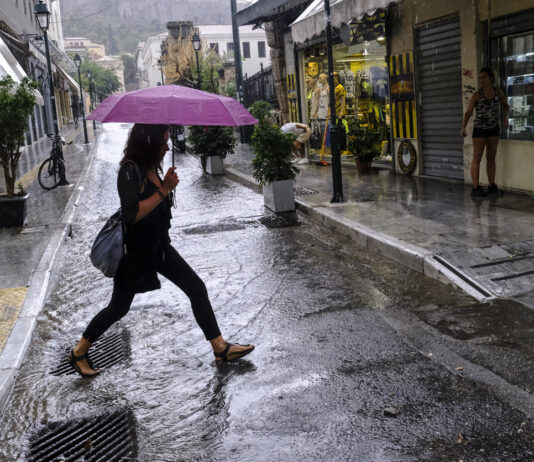 Frau mit Regenschirm überquert Straße im Regen. Im Hintergrund sieht man mehrere Geschäfte die hell erleuchtet sind.