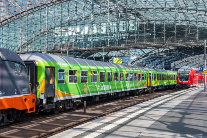 Ein grüner Flixtrain hält in einem Bahnhof. Daneben steht ein Zug der Deutschen Bahn. Die Sonne scheint durch das verglaste Dach des Bahnhofs.